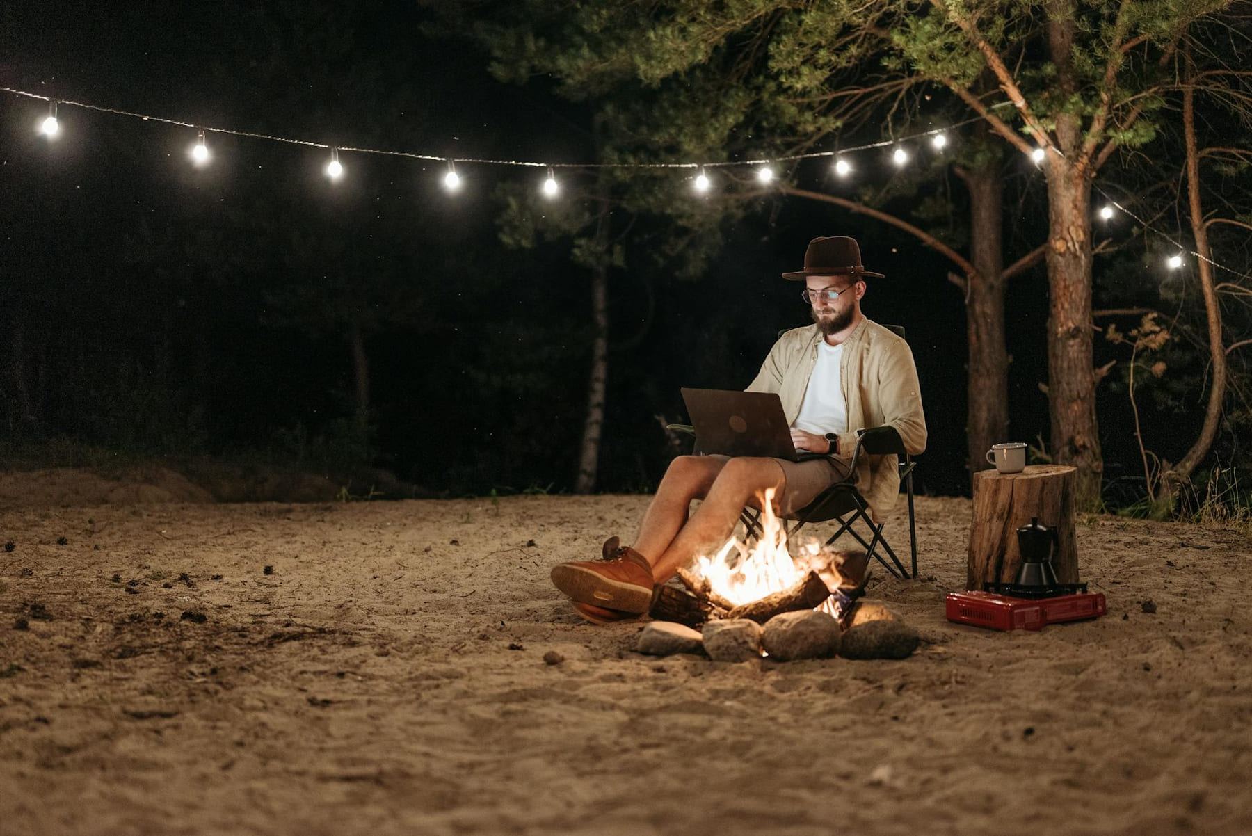 Digital nomad working remotely by a campfire with a laptop under string lights in the forest. on The Anywhere Office