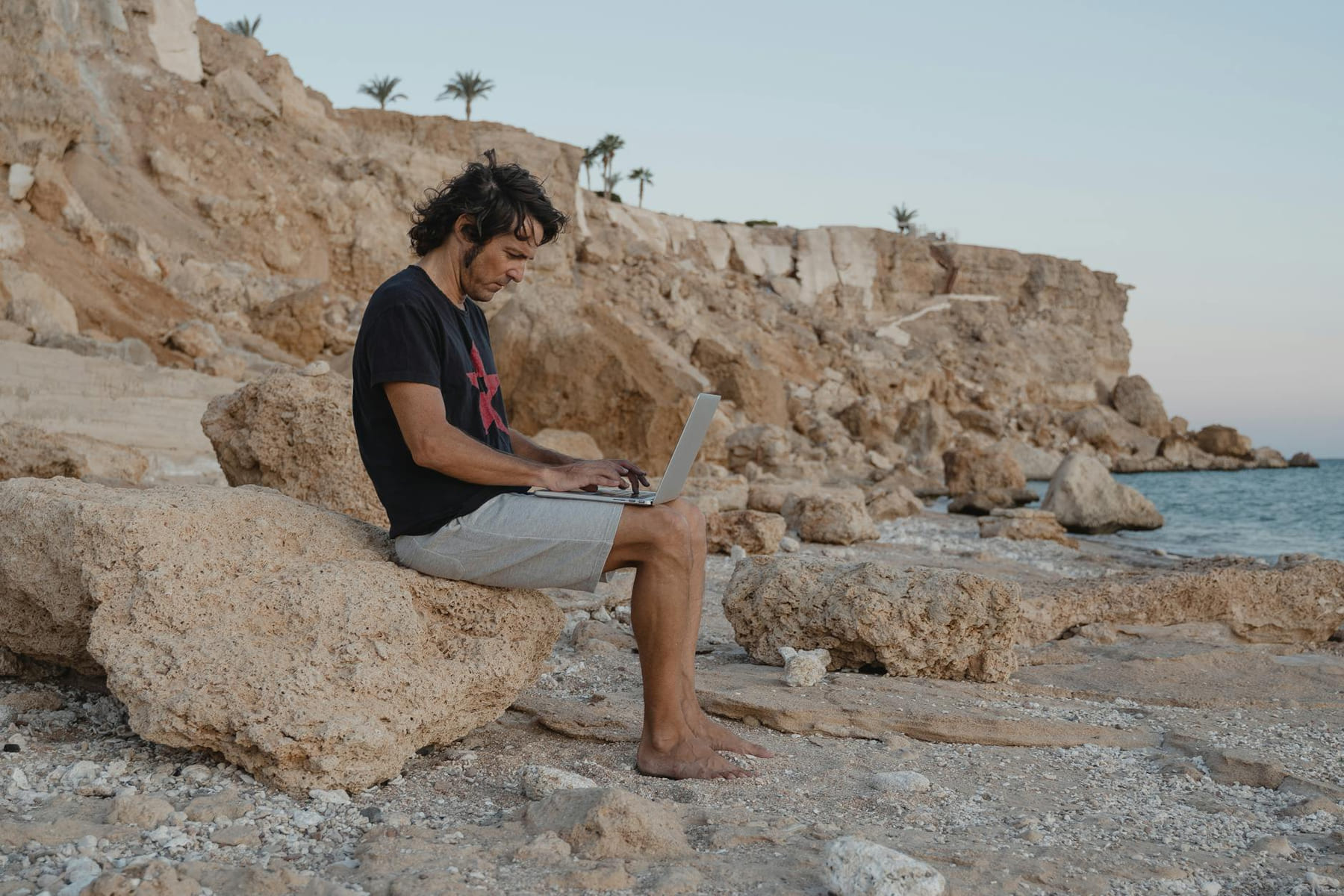 Man sits on rocks with laptop by the beach, enjoying remote work under a serene sunset. on The Anywhere Office