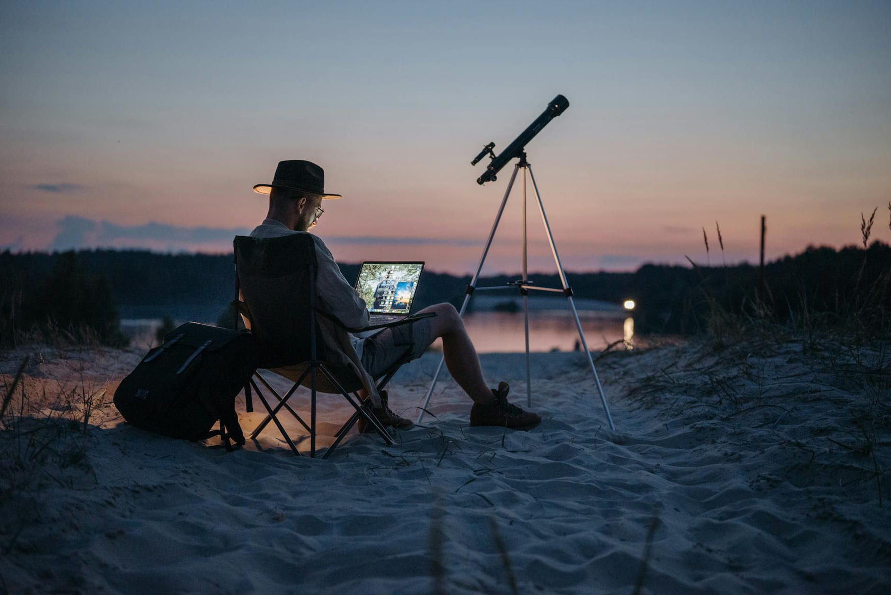 A man works on a laptop with a telescope nearby, on a sandy beach at twilight. on The Anywhere Office