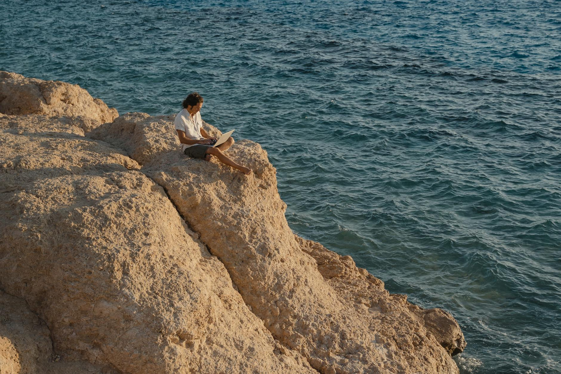 A man sits on a rocky cliff by the sea, working remotely on a laptop. on The Anywhere Office
