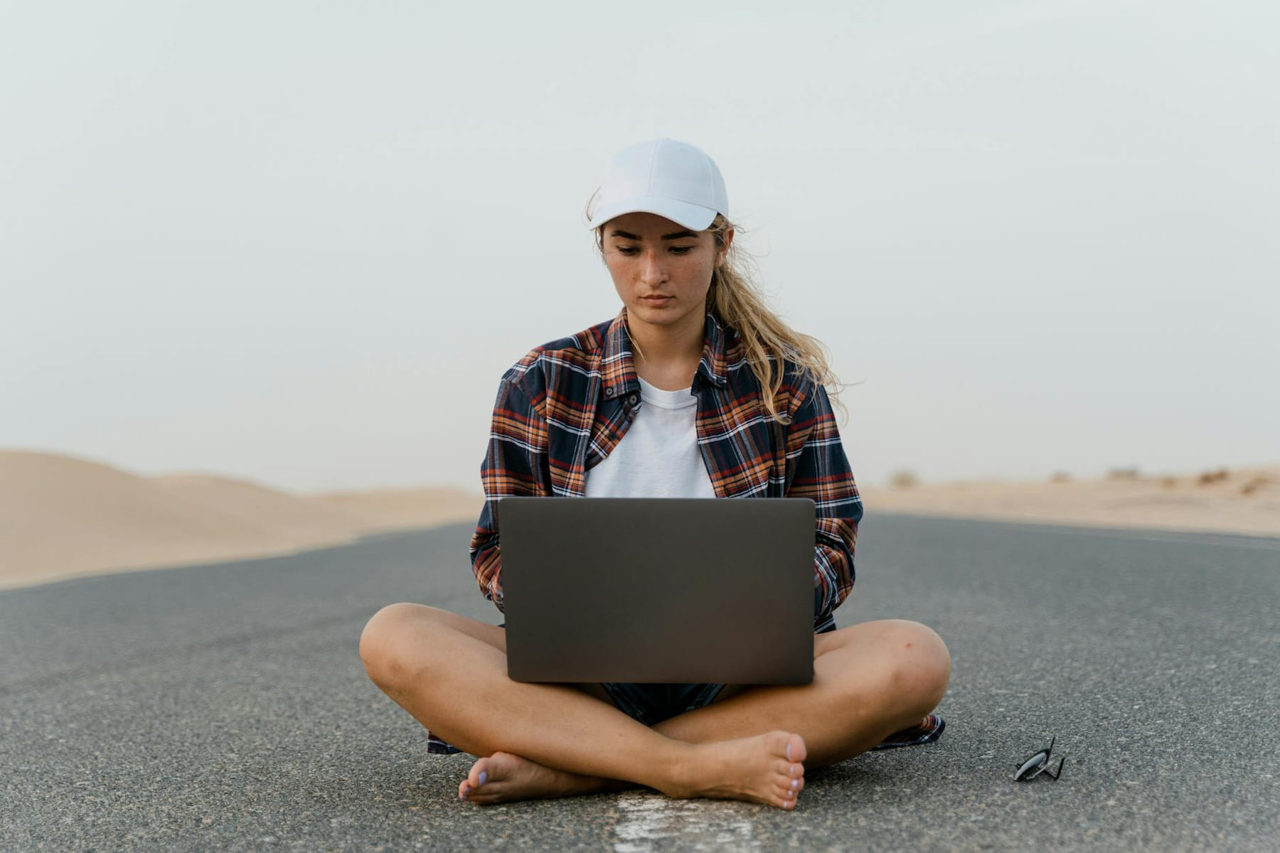 Focused young woman sits cross-legged on a road using a laptop, embodying remote work in a desert setting. on The Anywhere Office