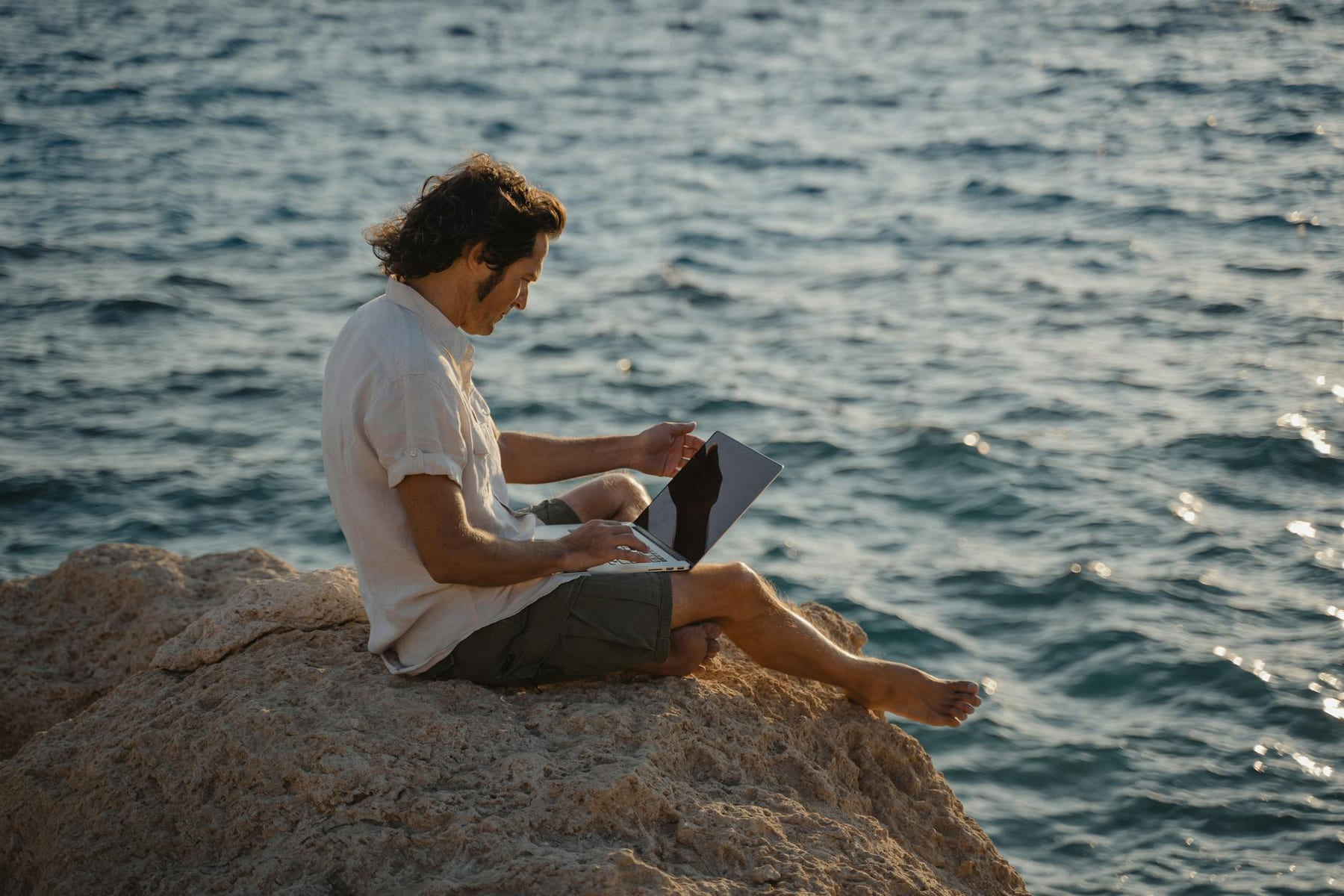A man sitting on rocky terrain by the sea, working remotely on his laptop during the day. on The Anywhere Office